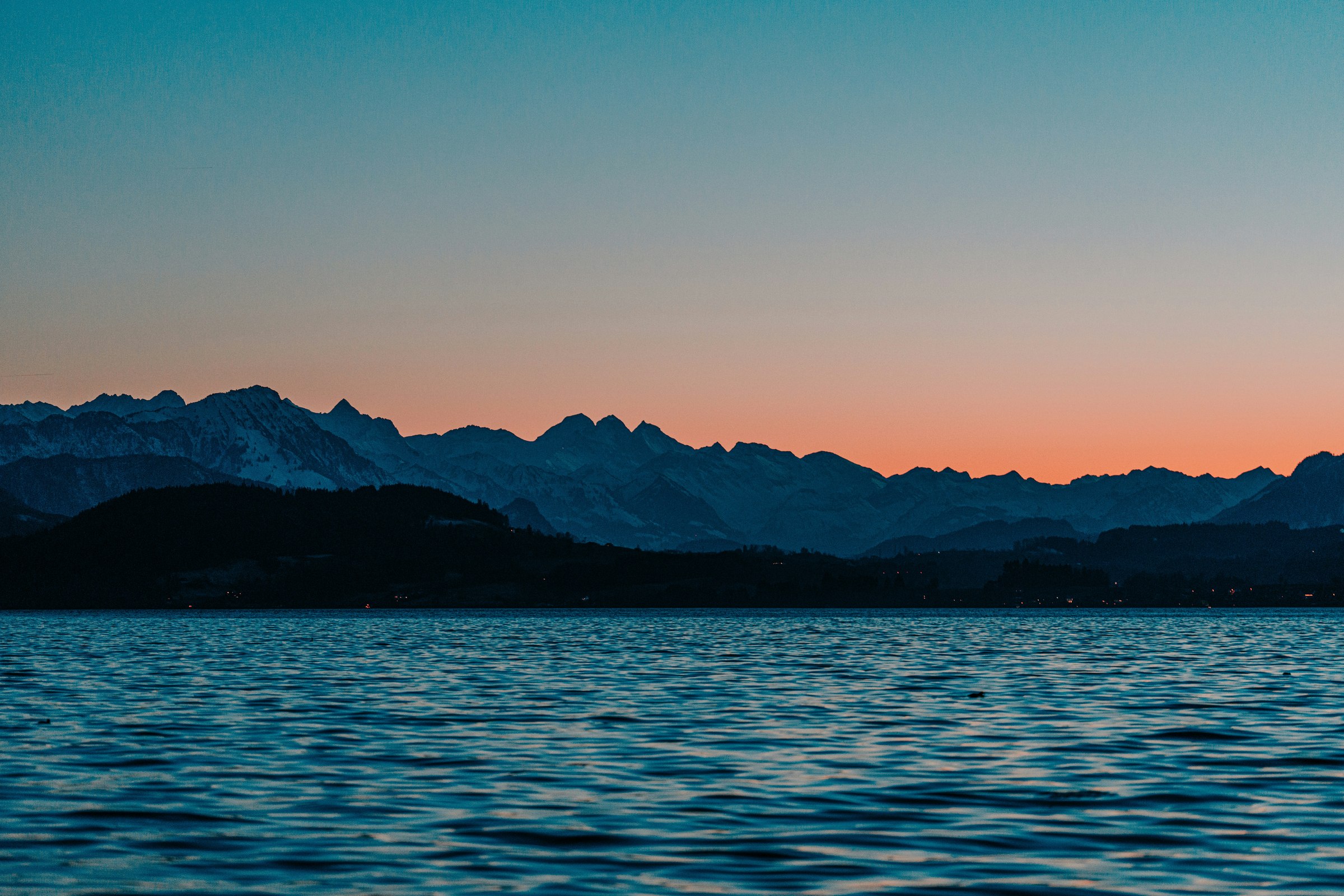 A deep blue Swiss lake at dusk below a dark mountain range and a band of orange sky
